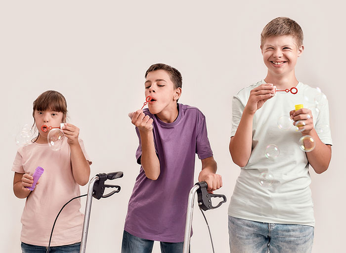 Three cheerful disabled children with Down syndrome and cerebral palsy smiling while blowing soap bubbles, standing together isolated over white background